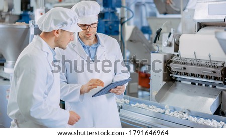 Two Young Male Quality Supervisors or Food Technicians are Inspecting the Automated Production at a Dumpling Food Factory. Employee Uses a Tablet Computer for Work. They Wear White Work Robes.