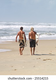 Two young male surfer dudes walking away from the camera along the beach carrying their surfboards with the waves crashing in the background.
