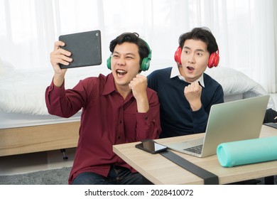 Two Young Man Friends Are Excited While Watching TV Online On Digital Tablet Stay At Home Have Fun. Couple Of Male Asian Happy Using Laptop Online Streaming Chatting Greeting People On Social Media.