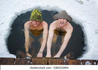 Two young man sitting in the cold water in ice-hole with snow around them after Russian bath on cold winter day
