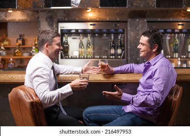 Two Young Man Talking At Counter. Two Friends Sitting In Bar And Drinking Whisky