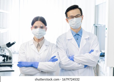 Two young masked intercultural scientists in whitecoats and gloves standing in front of camera while working over vaccine in laboratory - Shutterstock ID 1679724532