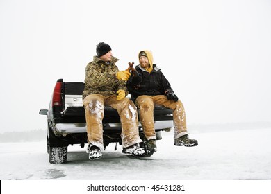 Two Young Men Enjoy A Beer While Sitting On The Tailgate Of A Truck In A Winter Environment. Horizontal Shot.