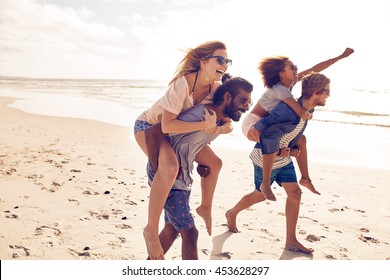 Two Young Men Giving Their Girlfriends Piggyback Rides At The Beach. Cheerful Young Friends Enjoying Summertime On The Beach.
