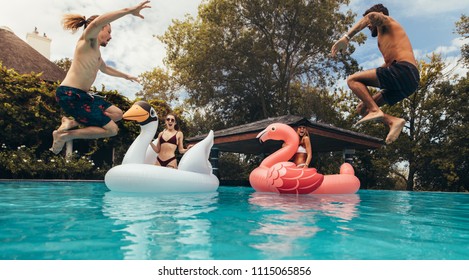 Two Young Men Jumping Into The Pool With Girls On Big Inflatable Toys. Group Of Young People Having Fun In A Swimming Pool.