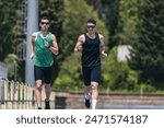 Two young men running in a park, one wearing a green tank top and the other wearing a black tank top