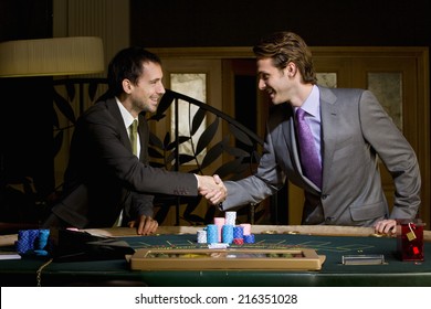 Two young men shaking hands over poker table, smiling, side view