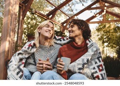 Two young men, smiling and enjoying each others company, cozy up under a colorful blanket, sipping warm drinks in a charming outdoor cafe filled with autumnal foliage. - Powered by Shutterstock