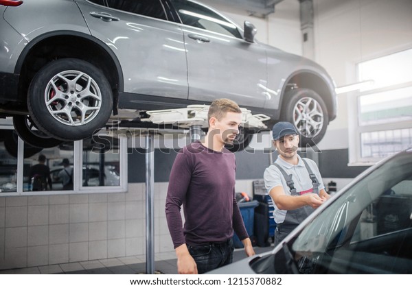 Two young men stand\
in garage at car. Worker points on automobile. Owner looks at it.\
They are serious and concentrated. There is another car on platform\
behind them.