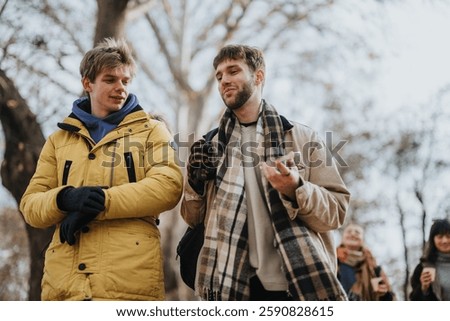 Two young men walking and conversing in an autumn park, enjoying camaraderie and relaxation. Trees and scarves suggest a chilly but pleasant atmosphere.