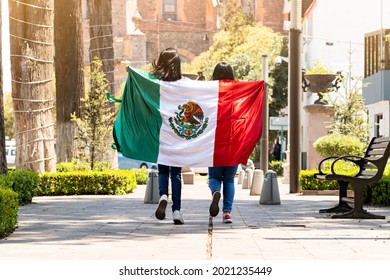 two young mexican women running on a walkway on a tree-lined street, with the mexican flag - Shutterstock ID 2021235449