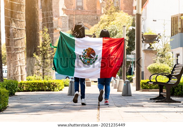 two young mexican women running on\
a walkway on a tree-lined street, with the mexican\
flag