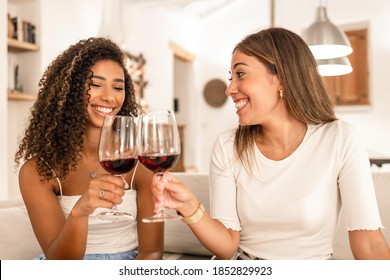 Two young mixed race woman celebrating at home toasting with red wine glasses - Smiling Hispanic curly brunette clinking with her best friend sitting at he sofa in her modern elegant living room