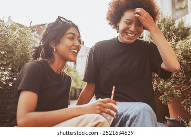 Two young mixed race women, one with curly hair, laughing together outdoors. Mixed race women enjoying a sunny day, women bonding and smiling, friends having fun. Diverse friends sitting outdoors. - Powered by Shutterstock