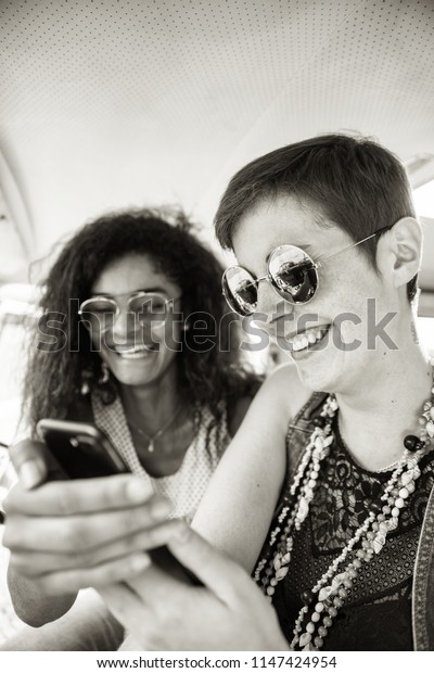 Two young\
mixed women having fun using a smartphone. They look vintage and\
going vacation in a van. black and\
white