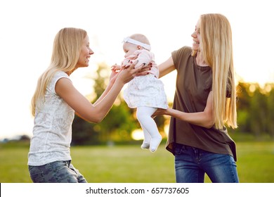 Two Young Mothers Are Walking In The Park In Summer With Her Baby. LGBT Lesbian Couple. The Concept Of  Family And Healthy Lifestyle