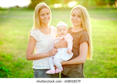 Two Young Mothers Are Walking In The Park In Summer With Her Baby. LGBT Lesbian Couple. The Concept Of  Family And Healthy Lifestyle