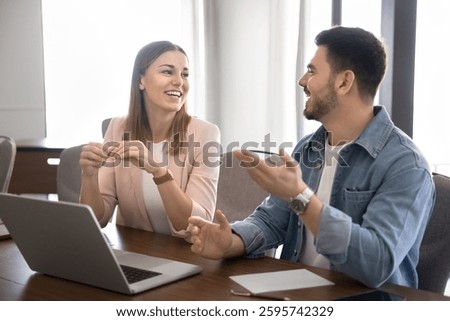 Two young multinational colleagues engaged in cheerful informal conversation, leading discussion sitting at desk with laptop in modern workspace, exchanging ideas for new project, joking and laughing
