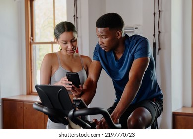 Two Young Multiracial Friends Using Mobile Phone On Exercise Bike At The Gym