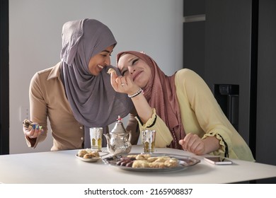 Two Young Muslim Women Laughing, Drinking Tea And Eating Arabic Sweets At The Kitchen Table.