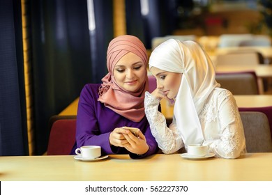 two young Muslim women sitting in a cafe and looking for something in a cell phone
