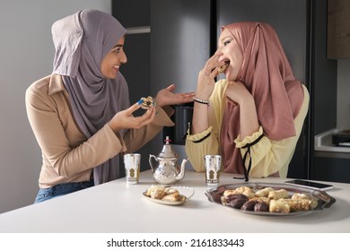 Two Young Muslim Women Talking, Drinking Tea And Eating Arabic Sweets At The Kitchen Table.