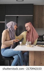 Two Young Muslim Women Talking, Drinking Tea And Eating Arabic Sweets At The Kitchen Table.