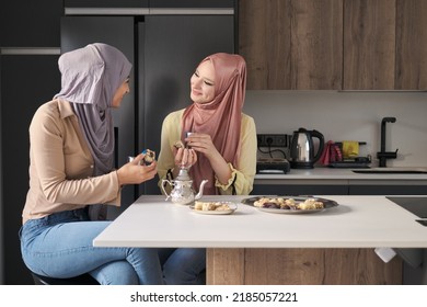 Two Young Muslim Women Talking, Drinking Tea And Eating Arabic Sweets At The Kitchen Table.