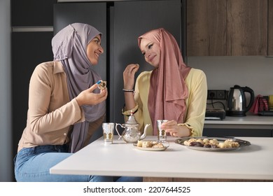 Two Young Muslim Women Talking, Drinking Tea And Eating Arabic Sweets At The Kitchen Table.