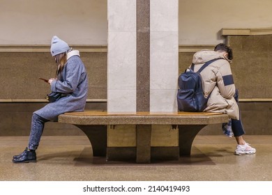 Two Young People, A Boy And A Girl, Are Sitting In The Lobby Of A Metro Station With Their Backs To Each Other. Both Of Them Are Passionate About Their Phones, Not Paying Attention To Anything Else.