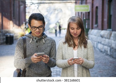 Two young people looking into smartphones while walking on city street