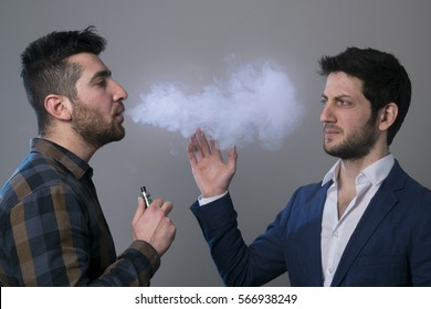 Two young people, one man is smoking or vaping an electronic cigarette and annoying his friend, vaping intruders. 