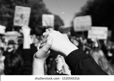 two young people at a rally, joining hands together signaling peace, unity and decisiveness in front of a crowd carrying protest placards with shallow depth of field, photographed in black and white