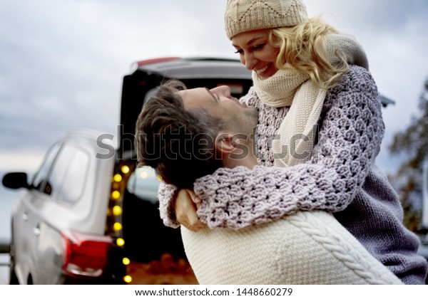 two young people\
in relationship hugging against the car and chrictmas lights.\
people wearing knitted\
clothes