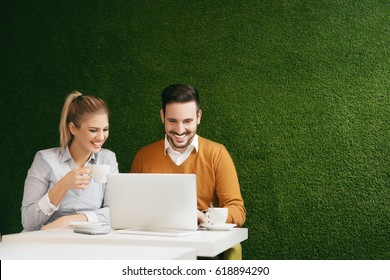 Two Young People Sitting Over A Green Grass Wall, Using A Laptop