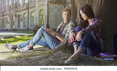 Two Young People Sitting Under Tree, Girl Looking Down And Guy Looking At Her