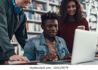 Deux jeunes gens souriants en regardant l'écran d'un ordinateur portable moderne et en aidant leur ami afro-américain à faire ses devoirs : photo de stock