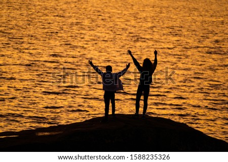 Two young people standing on rock in front of water with hands up looking at sundown. silhuette against sunset