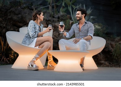 Two Young People Talking Sitting On Bright Armchairs In A Summer Night - Couple Of Young Adults Flirts During The Party In The Garden Of The Resort