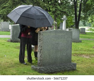 Two young people under an umbrella bringing a teddy bear and flowers to a grave site in the rain.