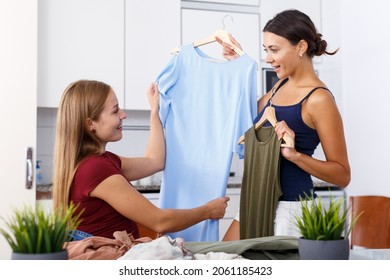Two young positive girlfriends looking new clothes at table and sharing in home