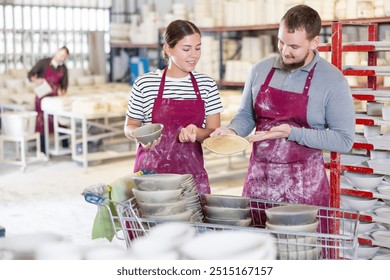 Two young pottery experts, girl and bearded man in maroon aprons, closely examining and discussing quality of handmade ceramics, surrounded by racks of pottery ware in sunlit studio - Powered by Shutterstock