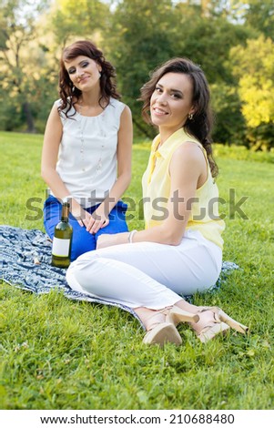 Two young pretty brunette girlfriends open a bottle of wine on the meadow during the picnic and sitting on the blanket. Vertical. Focus on the right girl.