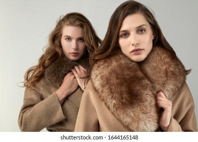 Two Young Pretty Models In A Luxurious Fury Brown Coats. Close Up Shot. White Background