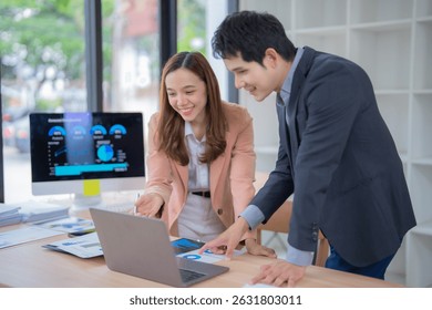Two young professionals engaged in a collaborative business discussion, analyzing data on a laptop in a modern office setting. A computer screen displays charts and graphs in the background
