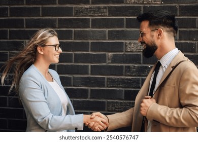 Two young professionals happily shaking hands in front of a rustic brick wall, sealing a successful partnership agreement. The urban scene exudes confidence and teamwork - Powered by Shutterstock