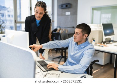 Two young professionals, one using a wheelchair, are working together on a computer project in a modern coworking office