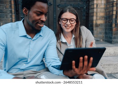 Two young professionals are using a tablet and smiling while sitting on steps in an urban environment