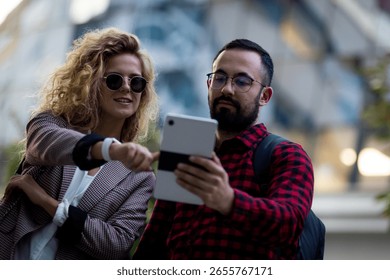 Two young professionals using a tablet in a city setting, engaging in discussion and sharing ideas. The man holds the device as the woman points, indicating collaboration and communication.