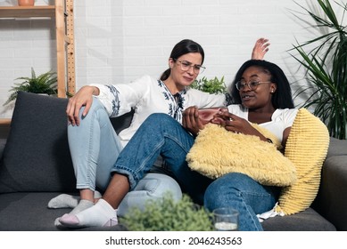 Two Young Roommate Woman Sitting On Sofa At Home And One Showing The Other Video On The Smartphone What She Did Last Night At The Crazy Party And She Feel Ashamed And Embarrassed